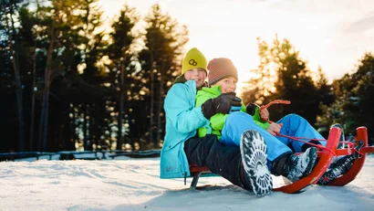 Zwei Kinder rodeln lachend im Schnee bei Sonnenuntergang