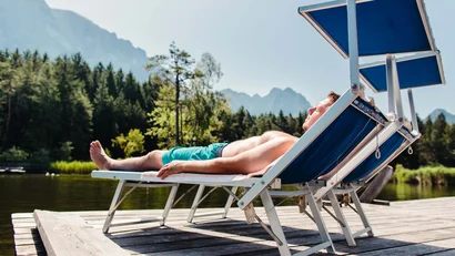 Mann entspannt auf Liegestuhl am See mit Wald und Bergen im Hintergrund