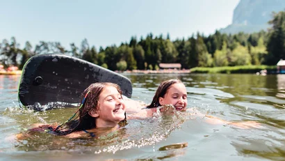 Zwei Mädchen schwimmen und lachen auf einem See mit Wald im Hintergrund