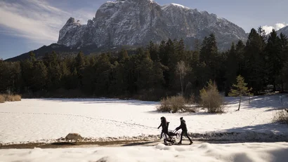 Zwei Personen mit Kinderwagen und Hund auf verschneitem Wanderweg vor Bergkulisse