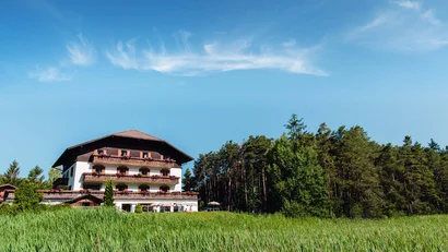 Mehrstöckiges Haus mit Balkon vor Wald und grünem Feld unter blauem Himmel