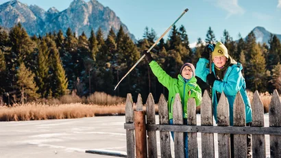 Zwei Kinder in Winterjacken an einem Holzzaun vor verschneiten Bergen