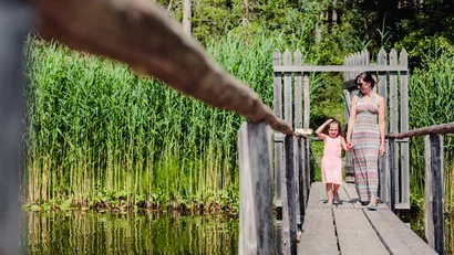 Frau und Kind gehen Händchen haltend über eine Holzbrücke am See