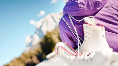 Person mit lila Jacke trägt weiße Schlittschuhe auf dem Rücken