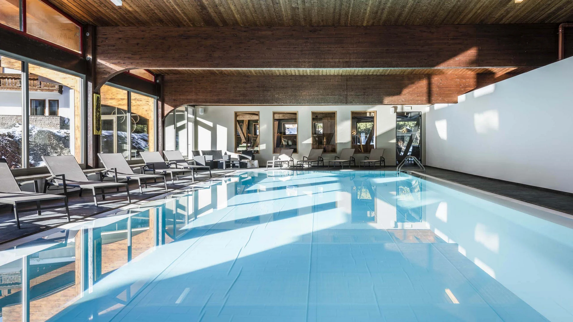Hallenbad mit Liegestühlen neben großen Fenstern und Holzbalkendecke