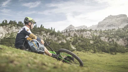 Radfahrer mit Helm sitzt auf Wiese und blickt auf Berglandschaft