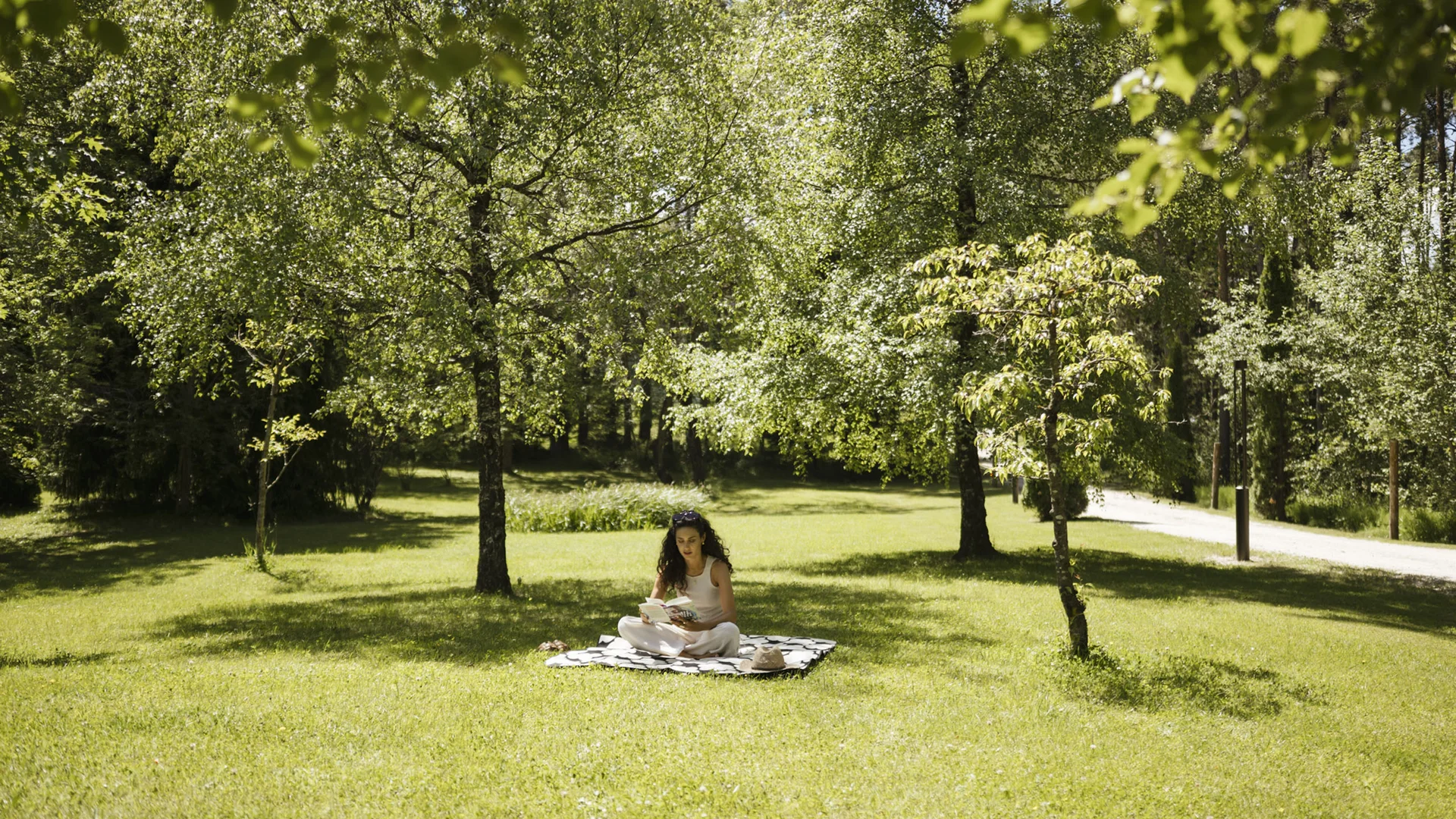 Woman sitting on blanket reading book on sunny grassy area with trees